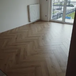 Nowe, jasnobrązowe deski ułożone w jodełkę w pokoju z białymi ścianami, grzejnikiem i dużym oknem balkonowym z widokiem na budynek mieszkalny.