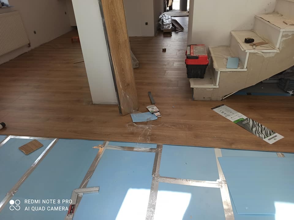 Fragment wnętrza w trakcie prac wykończeniowych, widoczne podłoże izolacyjne z folią aluminiową, częściowo położona podłoga, narzędzia i skrzynka narzędziowa obok schodów.