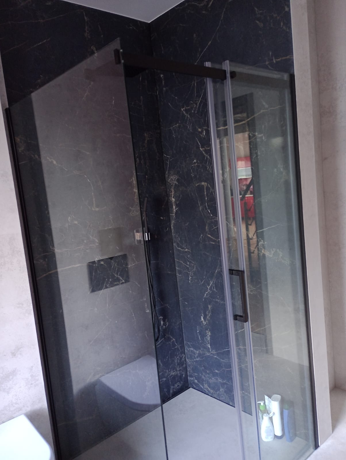 Nowoczesna kabina prysznicowa z czarnymi profilami i marmurowymi płytkami w odcieniach granatu i złota, widok z boku na otwarte drzwi i kosmetyki na podłodze.