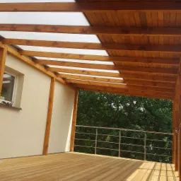 Zadaszony taras z drewnianą konstrukcją i przezroczystymi panelami dachowymi, widok z dołu na deski podłogowe i metalową balustradę.