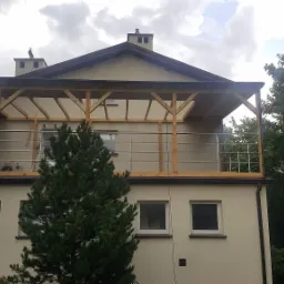 Drewniana konstrukcja z metalową balustradą na balkonie domu jednorodzinnego, widok z dołu, pochmurne niebo w tle.