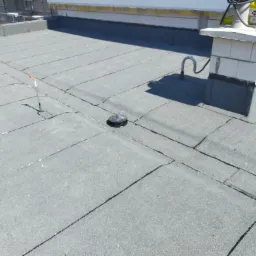 Szary, płaski dach pokryty papą z widocznym kominem z pustaków, przewodem wentylacyjnym i odpływem wody. Na kominie leżą paczki materiałów budowlanych.