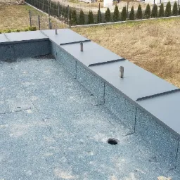 Płaski dach pokryty papą termozgrzewalną z metalową obróbką blacharską na krawędziach i pionowymi rurami wentylacyjnymi, widok z góry na tle trawnika i żywopłotu.