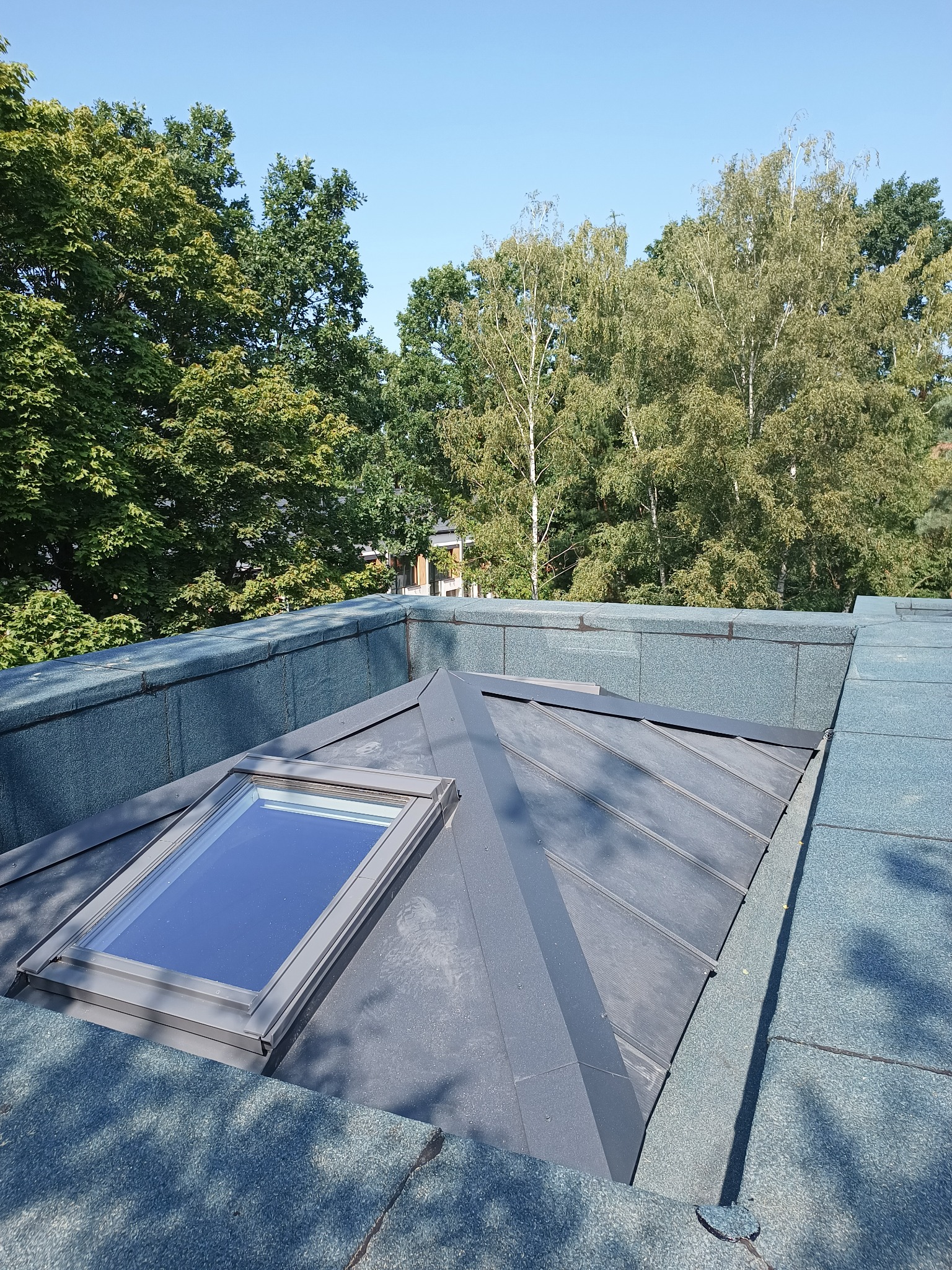 Widok z góry na fragment dachu z oknem połaciowym, pokrytego szarą papą, z zielonymi drzewami i błękitnym niebem w tle.