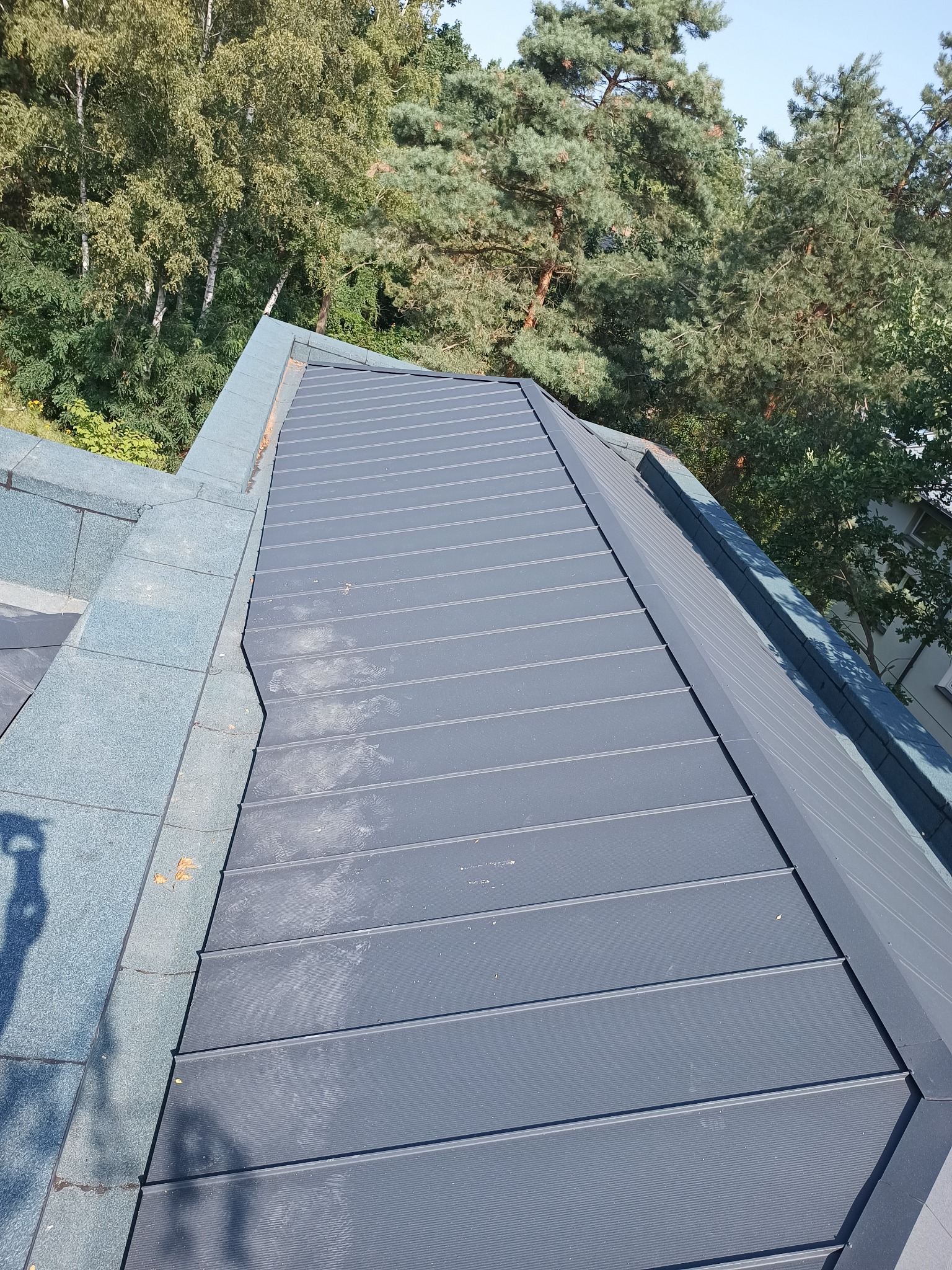 Szary dach z blachy na rąbek stojący, widok z góry, cień osoby robiącej zdjęcie widoczny na dachu, w tle drzewa.
