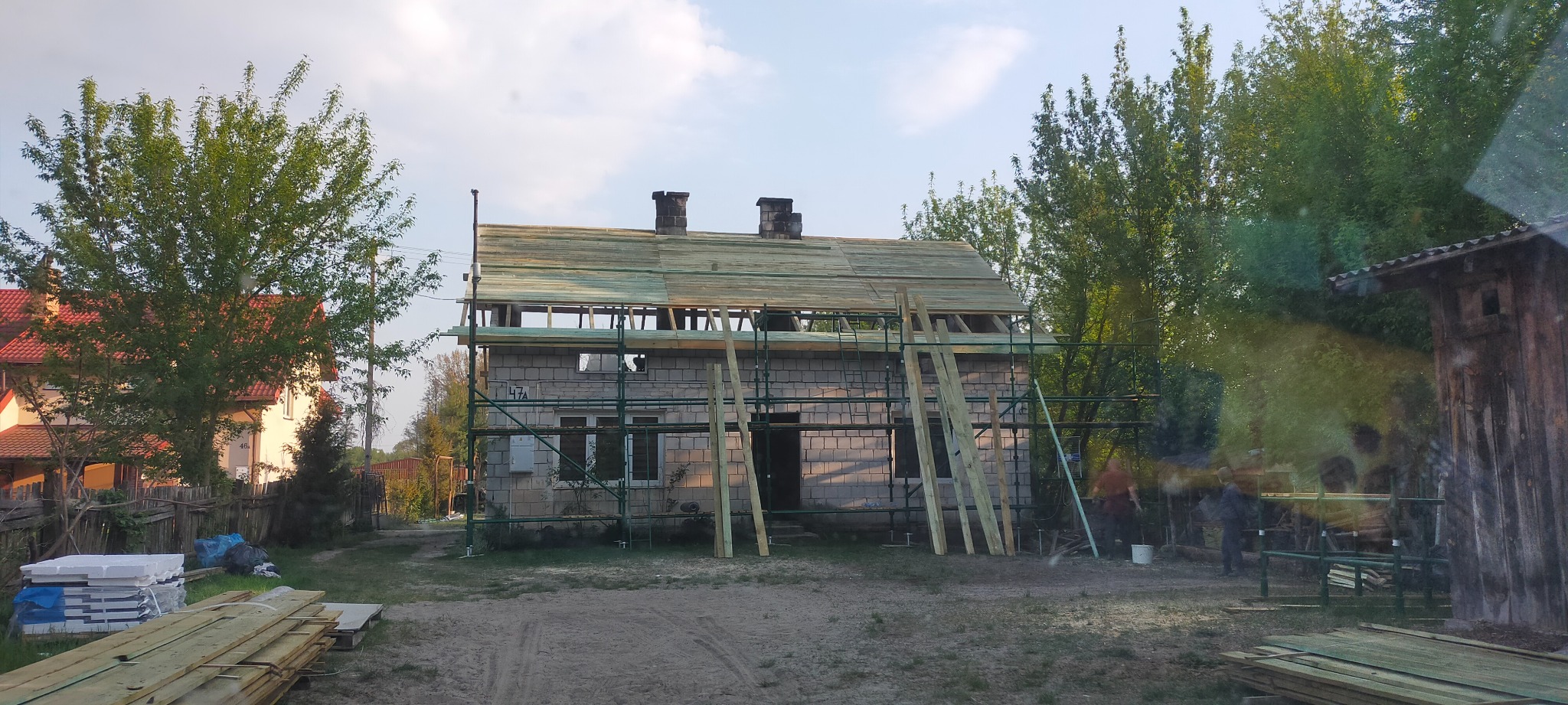 Budynek w trakcie budowy z widoczną konstrukcją dachu i rusztowaniami, stos drewna i materiałów budowlanych leżący na ziemi.