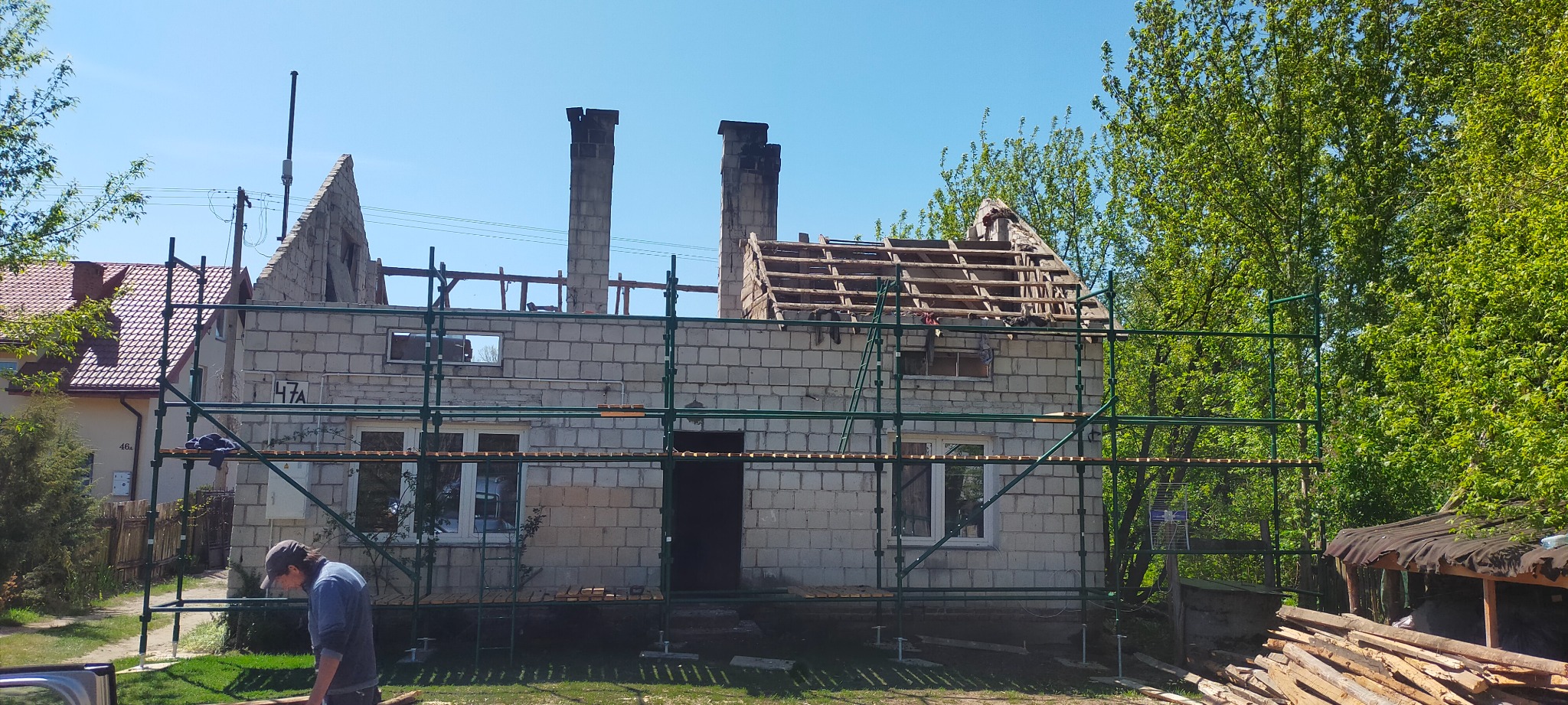 Budynek w trakcie remontu dachu, widoczne rusztowania, cegły, kominy i elementy konstrukcji drewnianej dachu. Na pierwszym planie mężczyzna w czapce i sterta drewna.