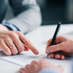 Podpis dokumentu: dłoń z pierścionkiem wskazuje tekst, druga dłoń podpisuje czarnym długopisem na białym stole.