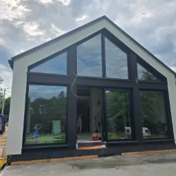 Nowoczesny dom z dużymi oknami w Gniezno, częściowo otynkowany na biało, z czarnymi ramami okiennymi. Na zewnątrz widoczny samochód dostawczy i materiały budowlane.