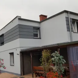 Dwukondygnacyjny dom z szarą elewacją, z wyraźnymi poziomymi pasami w górnej części, widok na zadaszony taras i fragment ogrodu z kwitnącym krzewem.