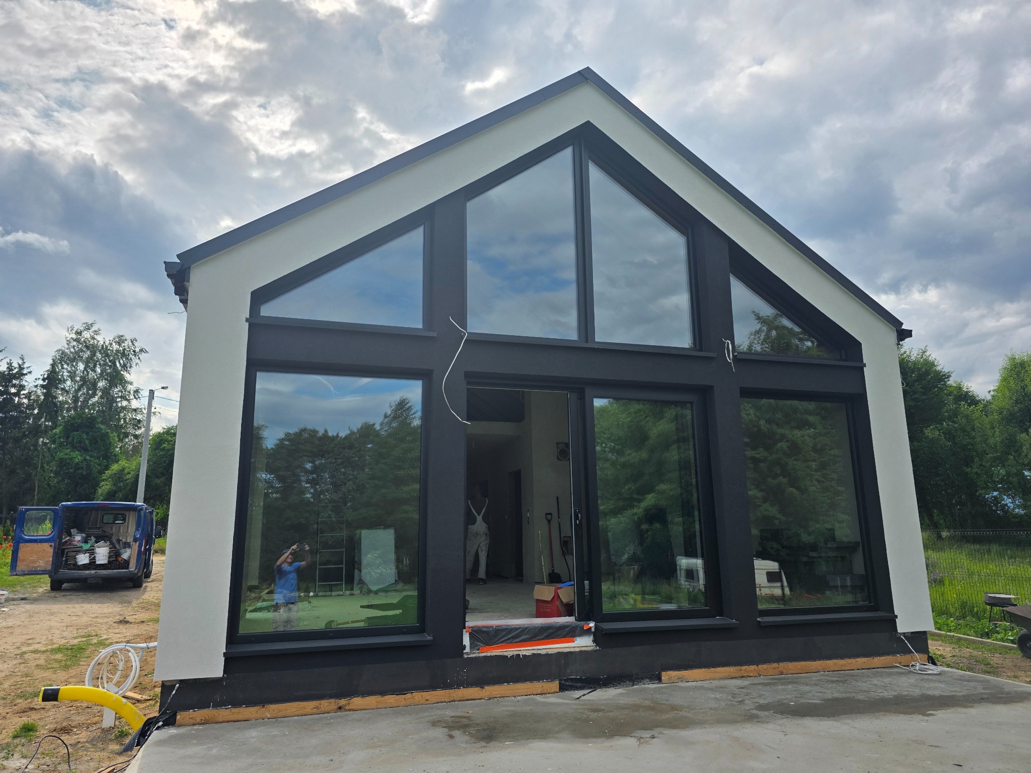 Nowoczesny dom z dużymi oknami w Gniezno, częściowo otynkowany na biało, z czarnymi ramami okiennymi. Na zewnątrz widoczny samochód dostawczy i materiały budowlane.