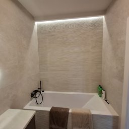 Pro-Home - Wykończona łazienka w beżowych płytkach z podświetlaną wnęką nad wanną. Minimalistyczna armatura i ręczniki w stonowanych barwach. Nowoczesne lustro z oświetleniem.