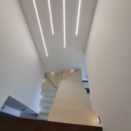 Pro-Home - Nowoczesne schody z oświetleniem LED w minimalistycznym wnętrzu. Białe ściany i sufit z wbudowanymi liniowymi lampami. Ciemna podłoga na dole schodów.