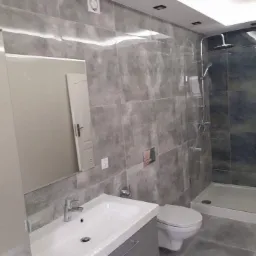 Nowoczesna łazienka z szarymi płytkami imitującymi beton, kabiną prysznicową typu walk-in i umywalką wpuszczoną w szarą szafkę, oświetlenie LED w suficie.