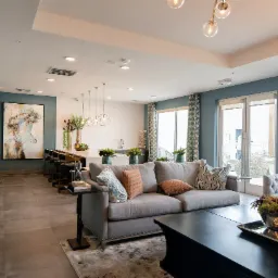 Przestronny salon z szarą sofą, czarnym stolikiem kawowym i otwartą kuchnią z hokerami, oświetlony nowoczesnymi lampami wiszącymi i obrazem na ścianie.