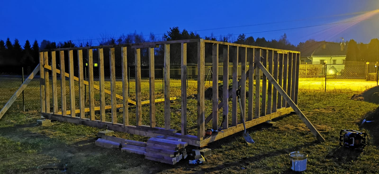 Wznoszenie drewnianej konstrukcji szkieletowej domu na fundamencie z bloczków betonowych, widoczne elementy mocujące i narzędzia, tło stanowią drzewa i oświetlony budynek w oddali, zdjęcie wykonane...