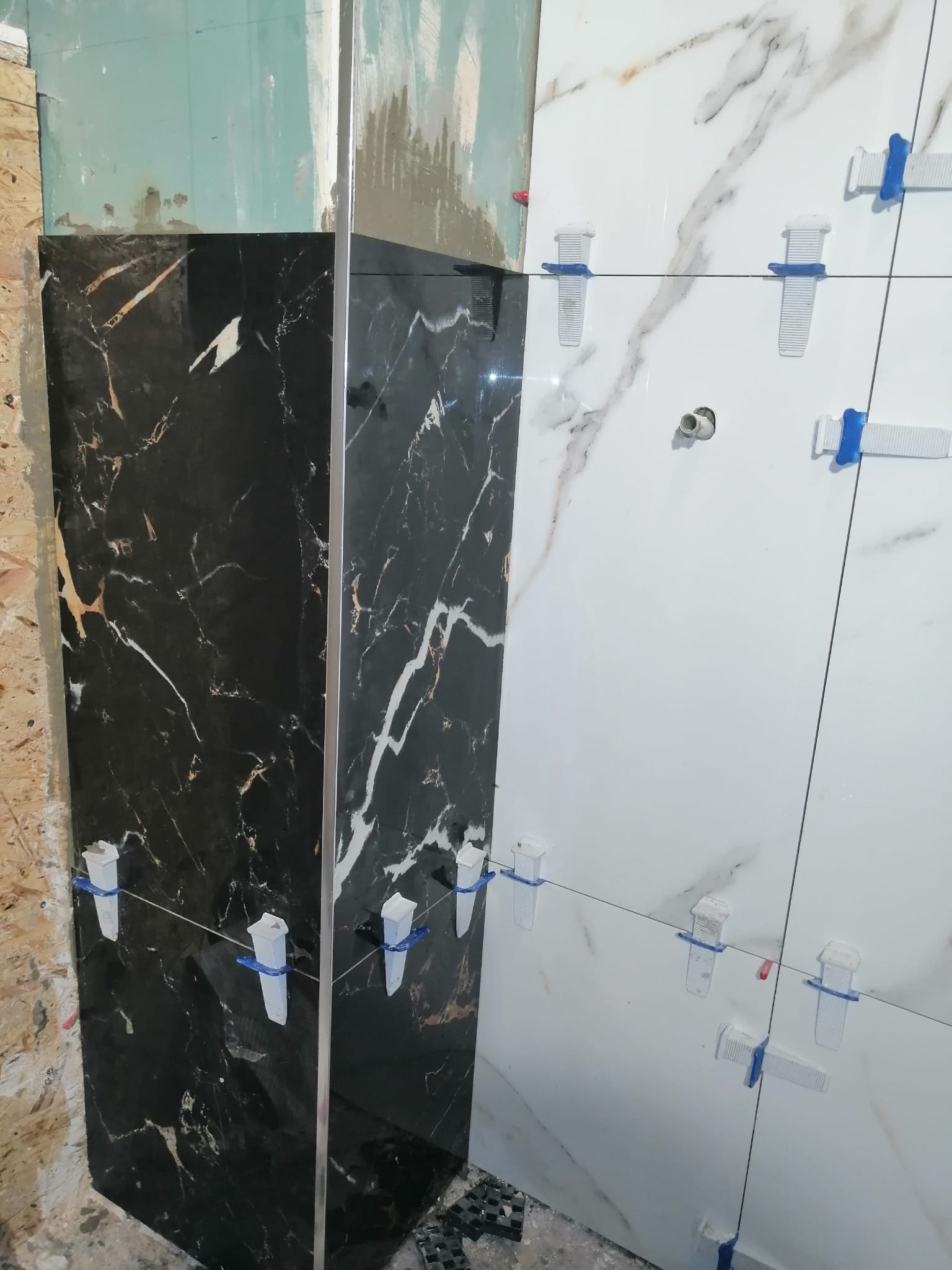 Układanie czarno-białych płytek imitujących marmur na ścianie, z użyciem systemów poziomowania, widoczne nierówne podłoże.