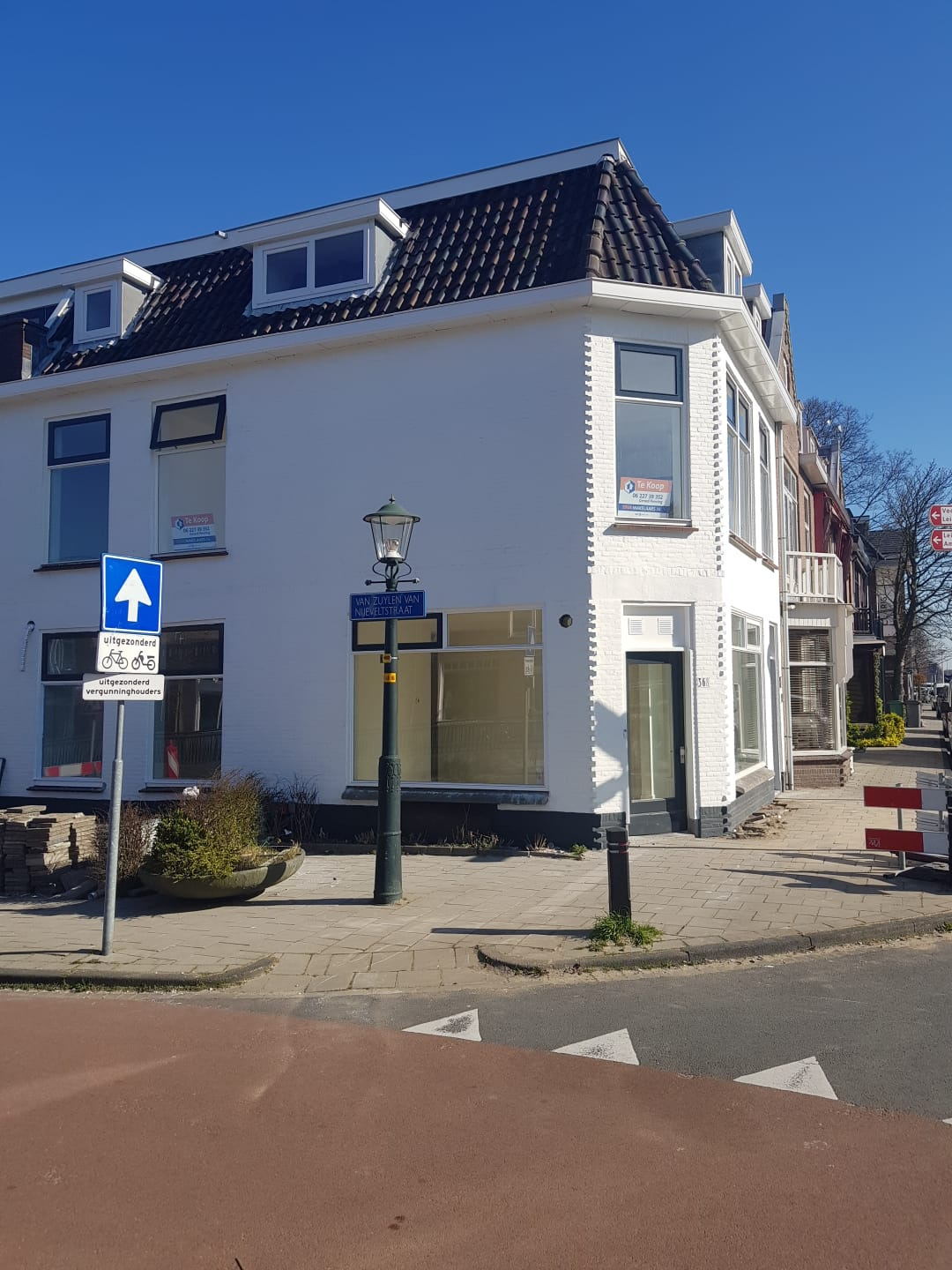 Biały budynek narożny z ciemnym dachem, widoczne okna z ogłoszeniami 'Te Koop', lampa uliczna i znak drogowy, ujęcie z poziomu ulicy w słoneczny dzień.