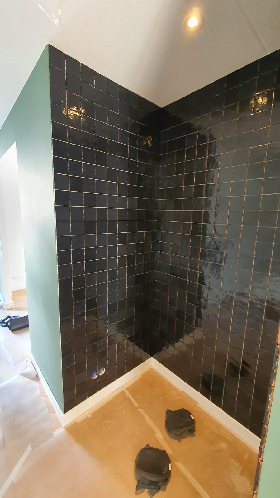 Ściana wyłożona błyszczącymi czarnymi płytkami w kwadratowym formacie z delikatną złotą fugą, widoczne gniazdka elektryczne, podłoga zabezpieczona folią malarską, fragment ściany pomalowany...