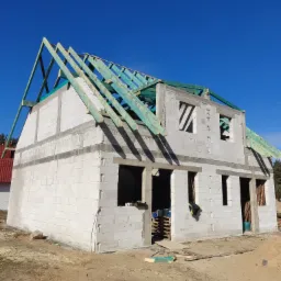 Surowy stan domu w trakcie budowy: ściany z betonu komórkowego, drewniana konstrukcja dachu pod gołym niebem, widoczne palety i resztki materiałów budowlanych.