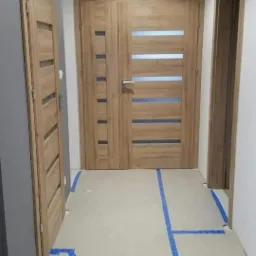 Montaż drzwi wewnętrznych z poziomymi przeszkleniami; widoczne zabezpieczenie podłogi niebieską taśmą malarską.