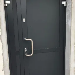 Ciemnoszare drzwi zewnętrzne z metalową klamką i srebrnymi zawiasami, zamontowane w surowej, betonowej futrynie.