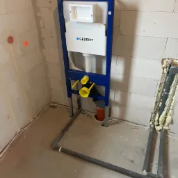 Stelaż podtynkowy Geberit zamontowany na ścianie z bloczków betonowych, widoczne rury doprowadzające wodę i odpływ kanalizacyjny w trakcie instalacji.