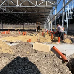 Robotnik zagęszcza ziemię wokół pomarańczowych rur kanalizacyjnych wewnątrz konstrukcji stalowej budynku, widoczny fragment wykopu i zbrojenia fundamentów.