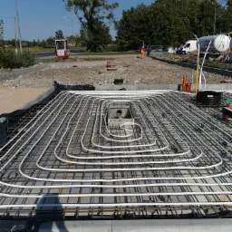 Przygotowanie podłoża pod wylewkę betonową z ułożonymi rurami ogrzewania podłogowego na siatce zbrojeniowej, widoczny zbiornik z gazem i minikoparka w tle.