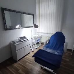 Gabinet zabiegowy z fotelem kosmetycznym przykrytym niebieskim pokrowcem i kocem, widoczne białe meble, lampa i lustro w ramie.