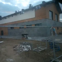 Budynek z elewacją częściowo wykończoną deską modrzewiową, kamieniem i jasnym tynkiem, widok zza ogrodzenia budowy, na ziemi gruz i materiały budowlane.