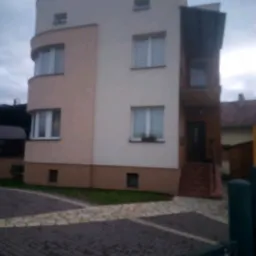 Trzypiętrowy dom z zaokrąglonym narożnikiem, pomalowany na beżowo i kremowo, z ciemnobrązową podmurówką i balkonem zadaszonym ciemną konstrukcją, widoczny z poziomu brukowanego podjazdu.