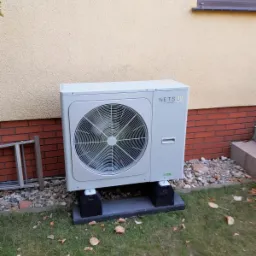 Zewnętrzna jednostka pompy ciepła Netsu, zamontowana na betonowych podstawach przy elewacji budynku z ceglaną podmurówką i żółtym tynkiem, widoczna drabina oparta o ścianę.