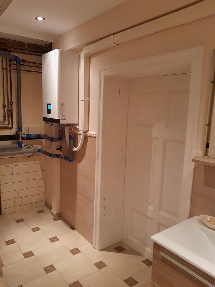 Biała pompa ciepła zamontowana na ścianie w pomieszczeniu z beżowymi płytkami i brązowymi akcentami, widoczne rury instalacyjne i białe drzwi.