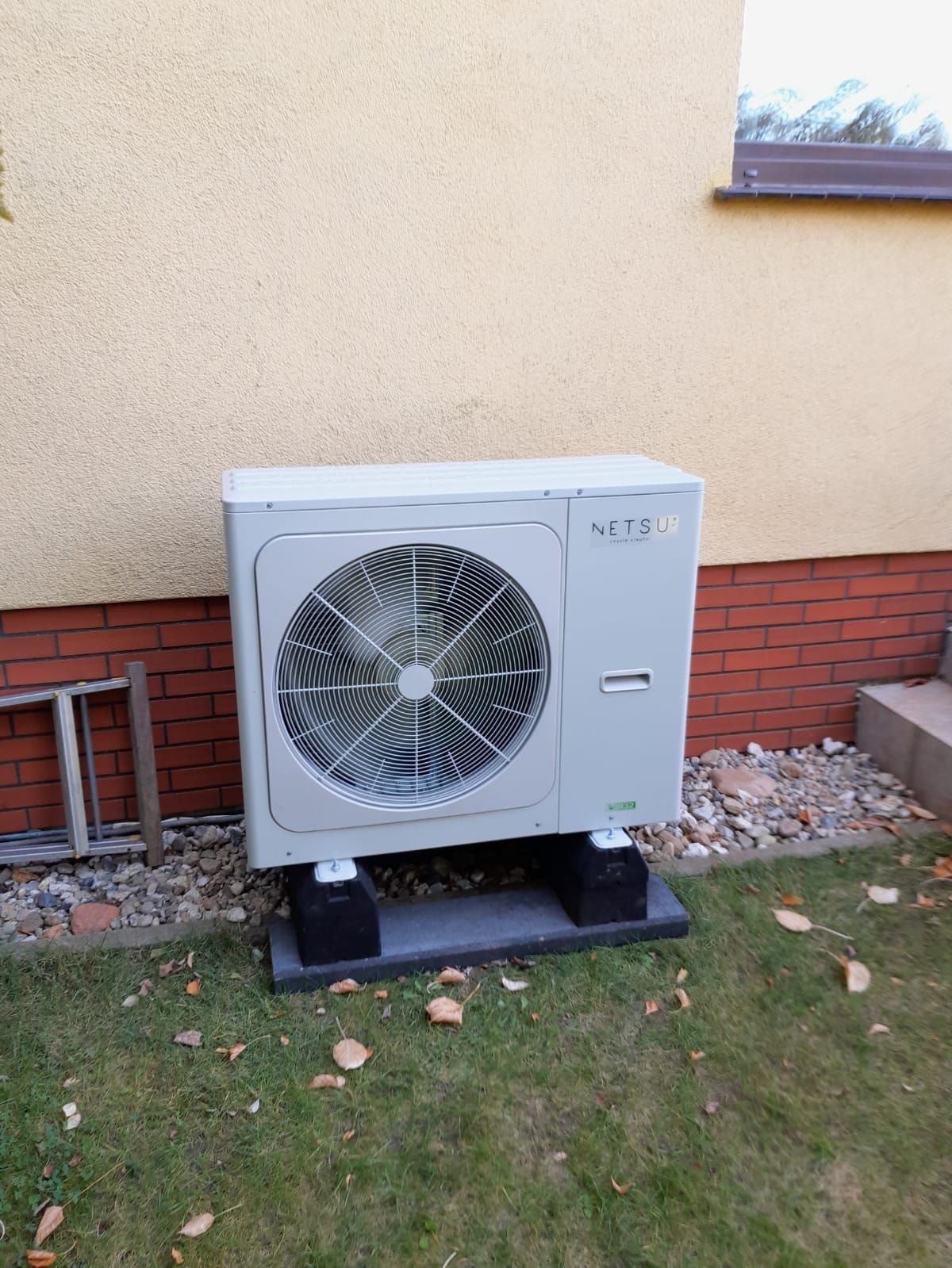 Zewnętrzna jednostka pompy ciepła Netsu, zamontowana na betonowych podstawach przy elewacji budynku z ceglaną podmurówką i żółtym tynkiem, widoczna drabina oparta o ścianę.
