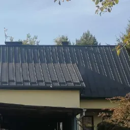 Czarny, blaszany dach trapezowy z anteną telewizyjną, zamontowany na jasnym, parterowym budynku z widocznymi rynnami i zielonym otoczeniem.