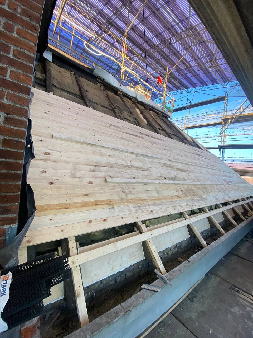 Wznoszenie drewnianej konstrukcji szkieletowej na fundamencie z cegły, widoczne rusztowania i zabezpieczenia nad głową.