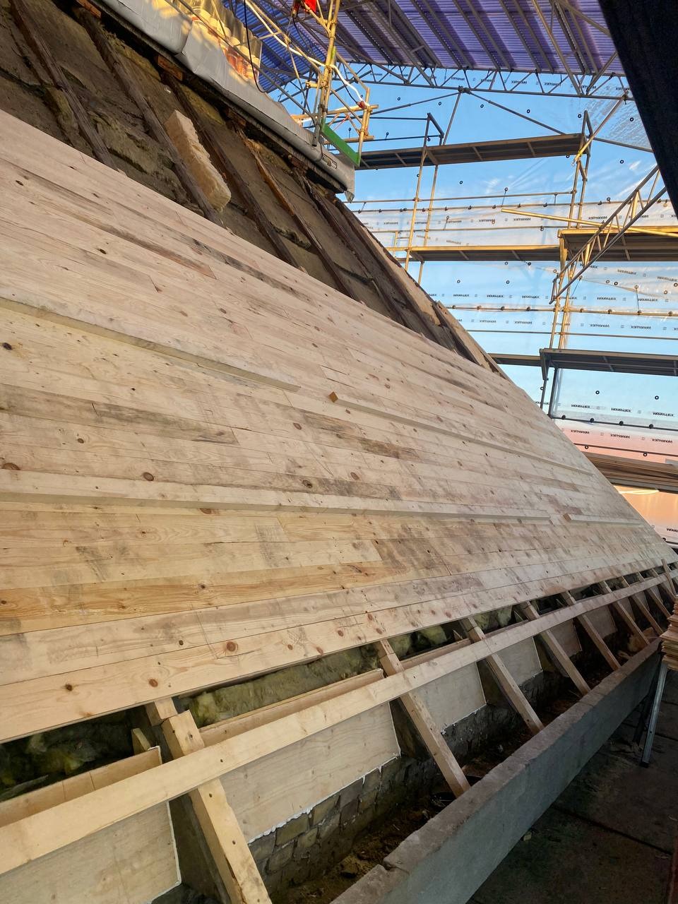 Konstrukcja drewnianego dachu w trakcie budowy, widoczne krokwie, izolacja i częściowo ułożone deski, rusztowanie w tle.