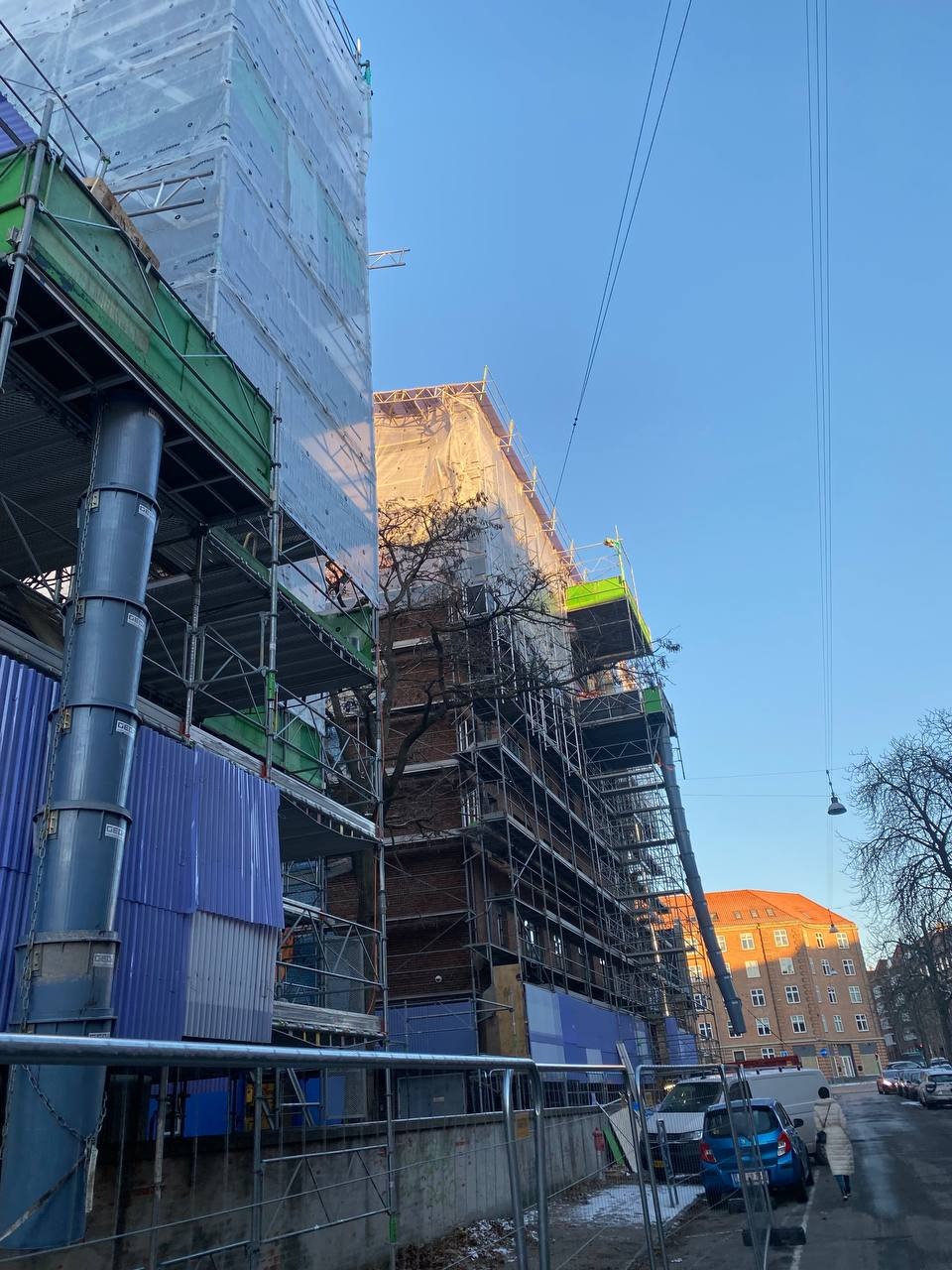Budynek w trakcie renowacji, otoczony rusztowaniami i częściowo przykryty folią ochronną, widoczny z perspektywy ulicy w słoneczny dzień.