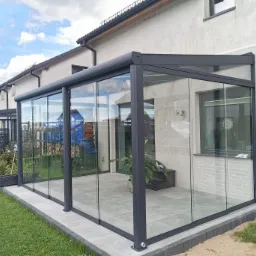 Szklana zabudowa tarasu z przesuwnymi drzwiami, szara konstrukcja, widok z zewnątrz, w tle dom jednorodzinny i błękitne niebo z chmurami.