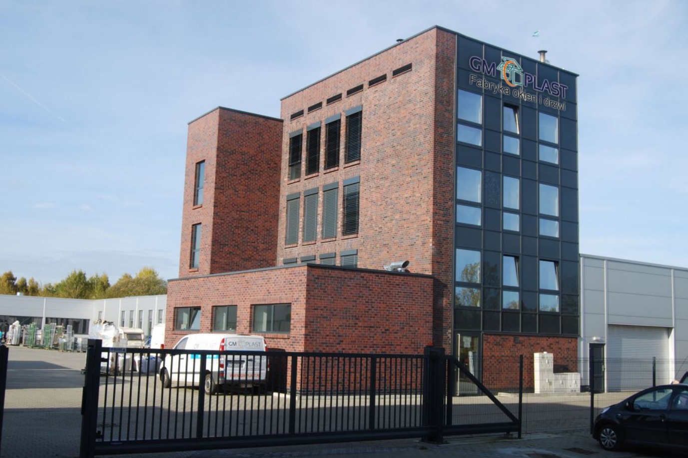 Budynek firmy produkującej okna i drzwi z cegły i paneli, z widocznym logo na szczycie, zaparkowane samochody i brama wjazdowa na pierwszym planie.