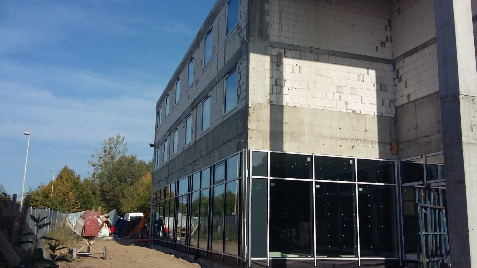 Budynek w trakcie budowy z zamontowanymi oknami aluminiowymi na parterze, widoczna surowa konstrukcja, ściany z betonu komórkowego i plac budowy z betoniarką.