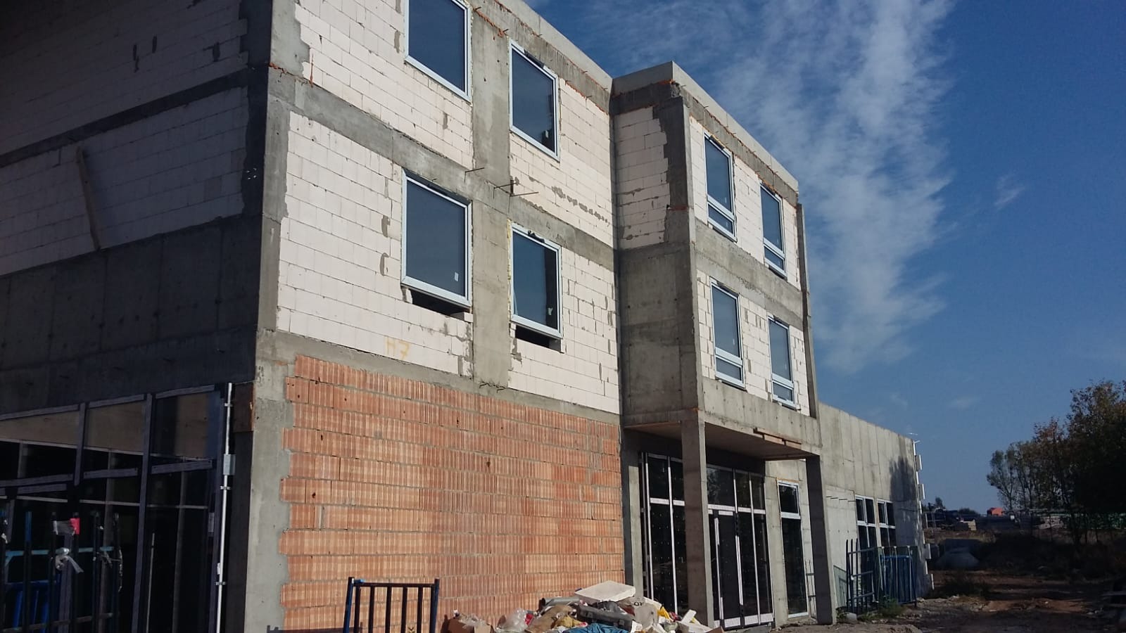 Budynek w trakcie budowy z zamontowanymi oknami z białymi ramami, widoczne materiały budowlane i nieporządek na placu budowy.