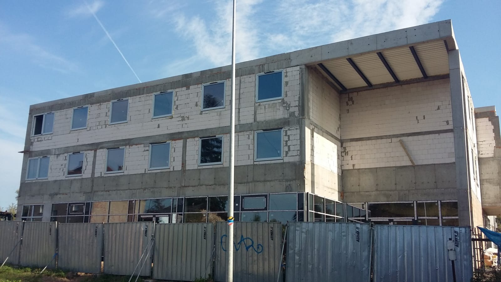 Budynek w trakcie budowy z zamontowanymi oknami PCV i aluminiowymi, widoczne surowe ściany z betonu komórkowego i metalowe ogrodzenie tymczasowe z niebieskim graffiti.