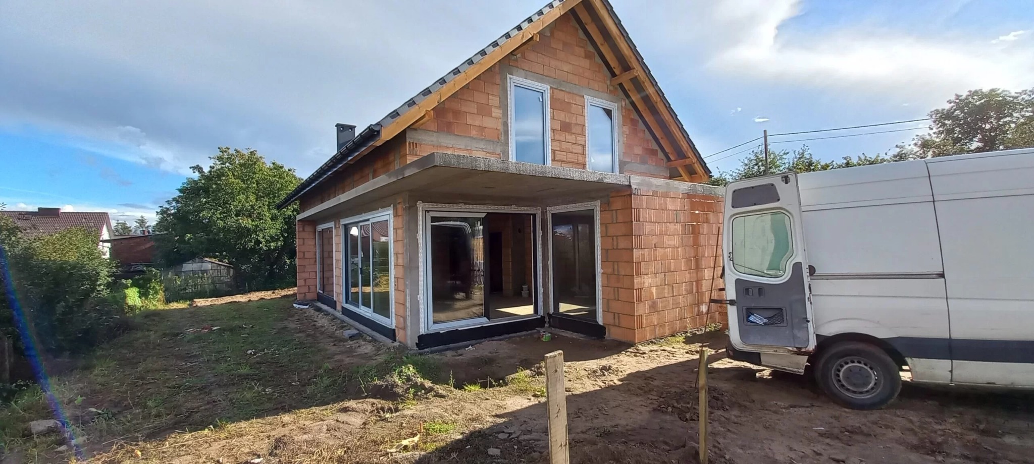 Dom w trakcie budowy z zamontowanymi oknami i drzwiami przesuwnymi, widoczny biały samochód dostawczy z otwartymi drzwiami.