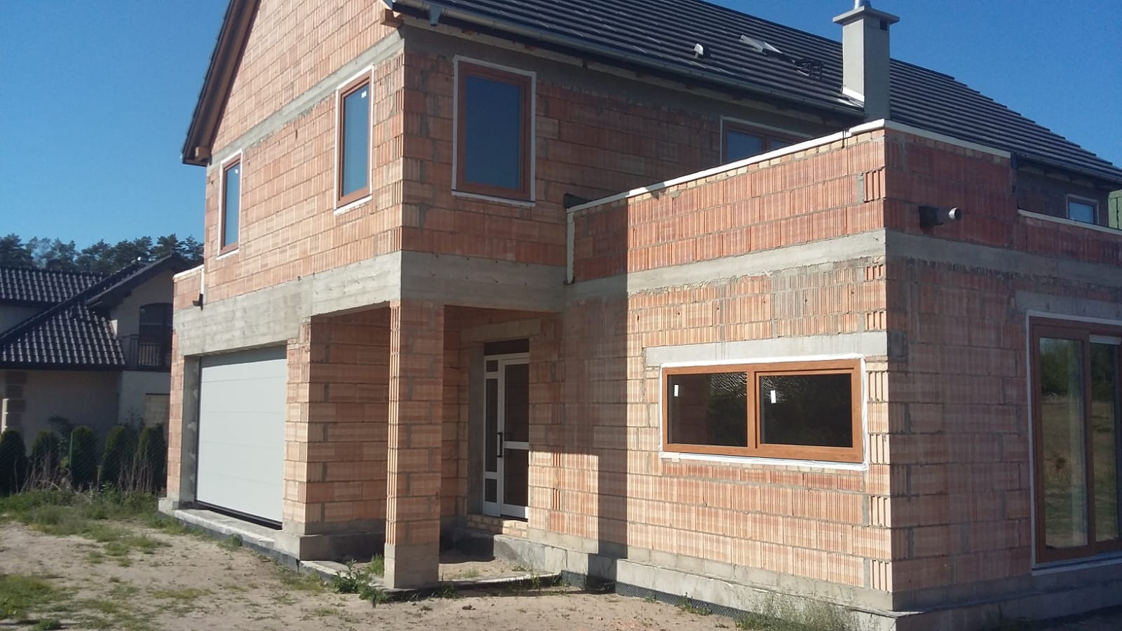 Dom w trakcie budowy z zamontowanymi oknami o drewnianych ramach i białymi drzwiami wejściowymi, widoczna brama garażowa.