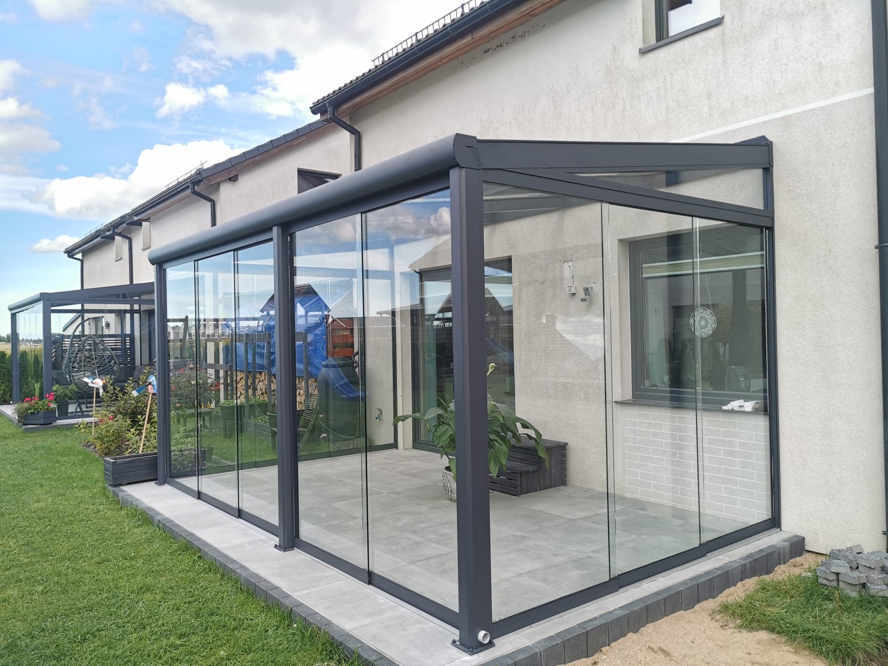 Szklana zabudowa tarasu z przesuwnymi drzwiami, szara konstrukcja, widok z zewnątrz, w tle dom jednorodzinny i błękitne niebo z chmurami.