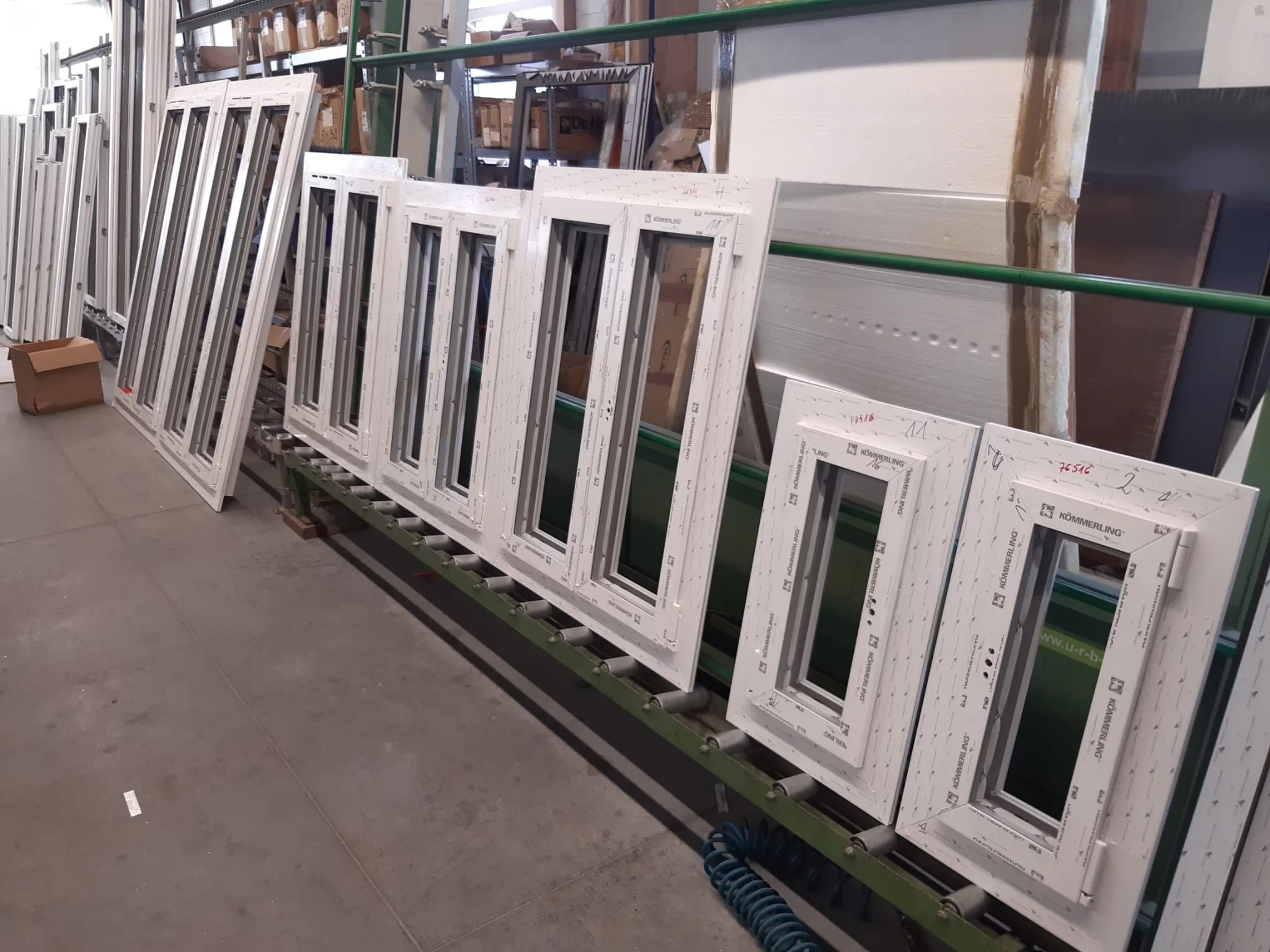 Białe ramy okienne marki Kömmerling ustawione w rzędzie na metalowym transporterze w hali produkcyjnej. Widoczne napisy i oznaczenia na profilach ram.