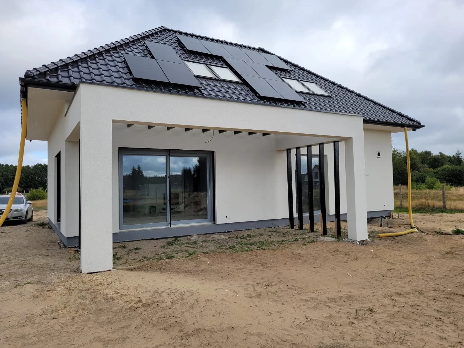 Nowoczesny dom jednorodzinny z panelami słonecznymi na dachu, dużym przesuwnym oknem tarasowym i minimalistyczną pergolą przy elewacji, widok z zewnątrz na plac budowy.