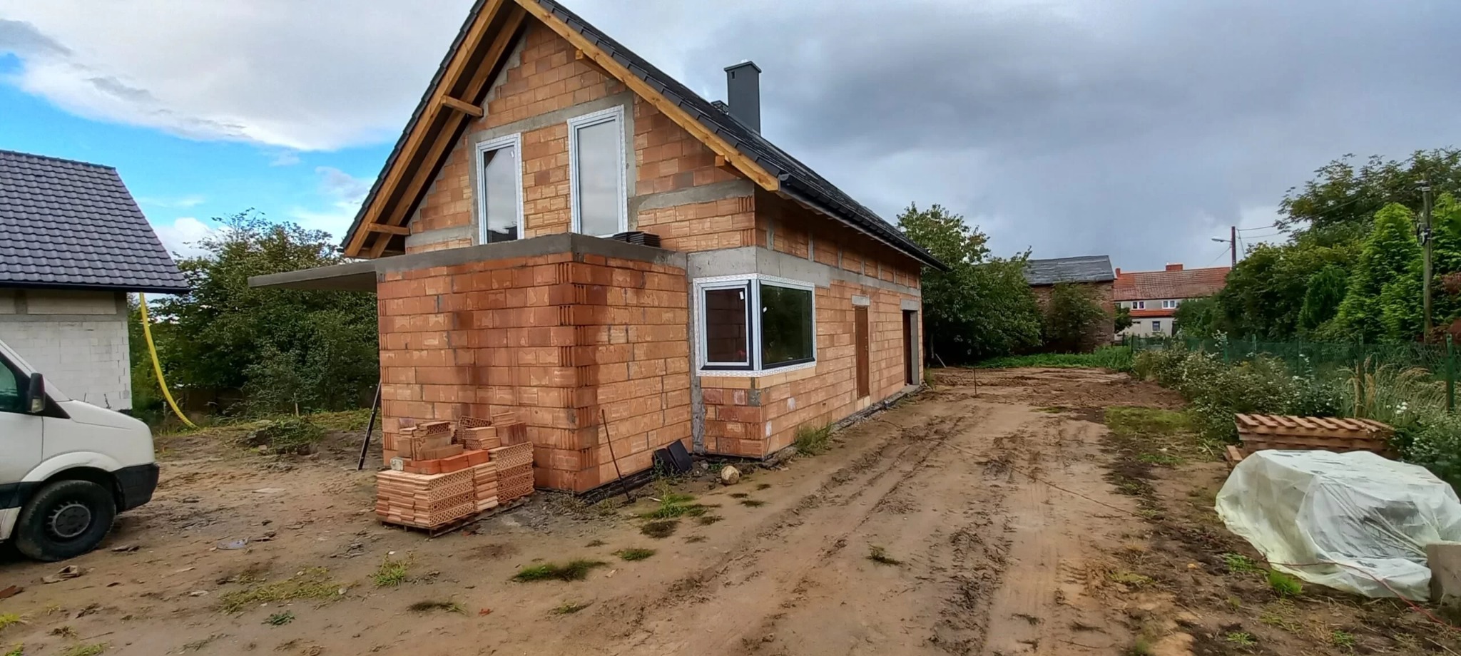Surowy budynek w trakcie budowy z zamontowanymi białymi oknami, widoczne cegły, dach pokryty dachówką, ziemia wokół budynku rozjeżdżona, pochmurne niebo w tle.